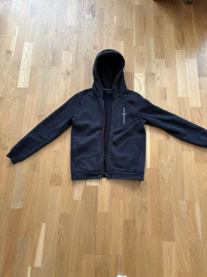 Svart hoodie från sail racing med zip - Snygg svart hoodie från sail racing i storlek 150 med dragkedja framtill och två fickor. Klassisk design med huva och diskret logga på bröstet. Perfekt för en avslappnad och sportig stil. Materialet känns mjukt och bekvämt.