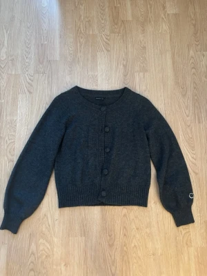 Mörkgrå kofta "Stockh lm" - Mysig mörkgrå kofta "Stockh lm" från MQ med rund halsringning och stora knappar framtill. Koftan har ballongärmar och ribbade muddar vid ärmslut och nederkant. Perfekt att slänga över en topp för en chill look.