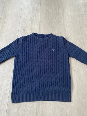Mörkblå kabelstickad tröja GANT - Snygg mörkblå kabelstickad tröja från GANT med rund halsringning och diskret broderad logga på bröstet. Tröjan har ribbade muddar vid ärmslut och nederkant, perfekt för en klassisk och stilren look. Materialet känns mjukt och bekvämt.