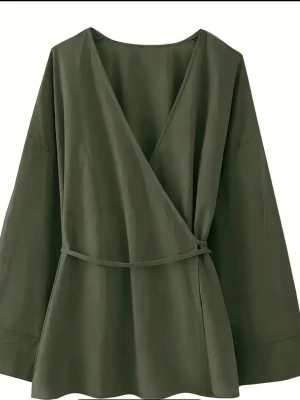 Olivgrön omlottblus med knytband - Stilren olivgrön omlottblus med långa ärmar och ett smalt knytband i midjan. Blusen har djup v-ringning och är tillverkad i ett mjukt, lätt tyg som ger ett snyggt fall. Perfekt för dig som gillar minimalistisk och trendig stil.