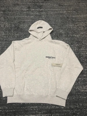 Grå hoodie från Fear of God Essentials - Snygg ljusgrå hoodie från Fear of God Essentials med svart logga på bröstet och stor text på ryggen. Klassisk passform med huva och ribbade muddar. Tillverkad i mjukt bomullsmaterial som känns riktigt skönt mot huden. Perfekt för en avslappnad streetwear-look.