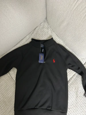 Svart ziptröja från Polo Ralph Lauren - Snygg svart ziptröja från Polo Ralph Lauren med klassisk röd logga broderad på bröstet. Tröjan har halv dragkedja, ribbade muddar och står upp lite i kragen. Perfekt för en stilren och avslappnad look.
