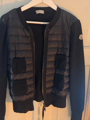 Svart cardigan  från Moncler - Snygg svart cardigan jacka från Moncler med quiltad front och fyra fickor. Jackan har långa ärmar i stickad kvalitet och dragkedja framtill. Moncler-logga på ärmen och ribbade muddar. Perfekt för lager-på-lager och streetstyle.