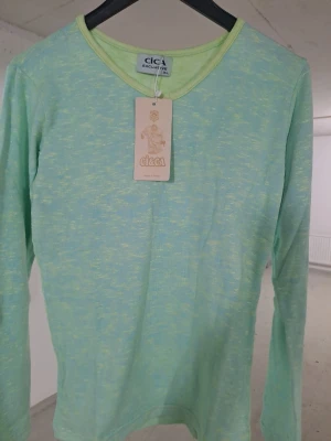 Grönmelerad långärmad tröja Cica - Fräsch långärmad tröja från Cica i grönmelerat tyg med V-ringning och mjuk känsla. Perfekt för dig som gillar en avslappnad och stilren look. Tröjan har en enkel design och passar till många olika outfits.