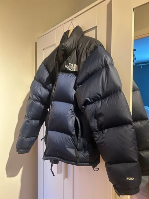 Marinblå vinterjacka The North Face Nuptse 700 - Säljer en klassisk marinblåpufferjacka från The North Face, modell 700. Jackan har hög krage, dragkedja framtill, justerbar nederkant och logga på bröstet och ryggen. Perfekt för kalla dagar med sin quiltade desig.