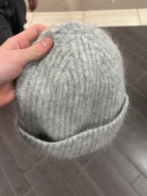 Ljusgrå stickad mössa - Mysig ljusgrå stickad mössa med bred uppvikt kant. Tillverkad i ett fluffigt, tjockt garn som ger en avslappnad och trendig look. Perfekt för att hålla värmen under kalla dagar och passar till de flesta outfits.