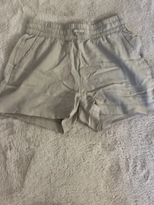 Beige shorts med resårmidja - Snygga beige shorts med resår i midjan och sidofickor. De har en avslappnad passform och är tillverkade i ett mjukt linnematerial, perfekta för varma dagar. En bakficka ger extra detalj. Passar dig som gillar enkel och stilren look.