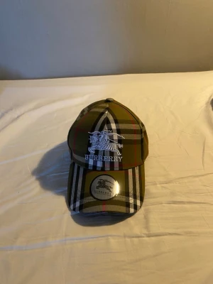 Burberry keps med klassiskt rutmönster - Säljer en snygg Burberry keps med det ikoniska rutmönstret i olivgrönt, svart, vitt och rött. Kepsen har broderad Burberry-logga och riddare framtill samt justerbart spänne bak. Perfekt accessoar för dig som vill sticka ut med stil.