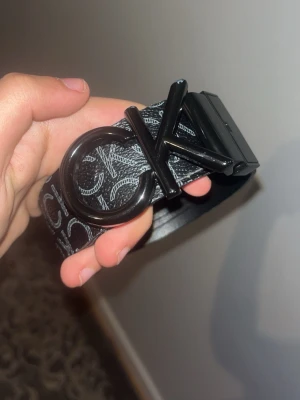 Äkta Svart Calvin Klein Reversibel Bälte - Snyggt svart bälte från Calvin Klein med stort CK-logotypspänne i metall. Bältet har ett mönster med vita CK-loggor längs hela remmen och är gjort i konstläder. Perfekt accessoar för att lyfta din outfit.