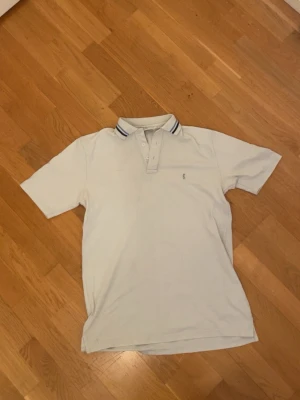 Vit pikétröja från Yves Saint Laurent - Klassisk vit pikétröja från Yves Saint Laurent med krage och knappar framtill. Tröjan har diskreta blå ränder på kragen och är kortärmad. Perfekt för en clean och stilren look.