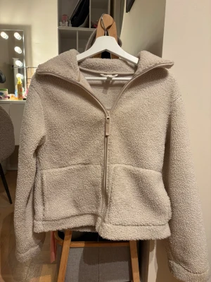 Beige teddyjacka med dragkedja - Mysig beige teddyjacka med huva och hel dragkedja framtill. Jackan har två stora fickor och är i en loose passform, perfekt för att hålla värmen. Tillverkad i mjukt syntetmaterial som känns skönt mot huden.