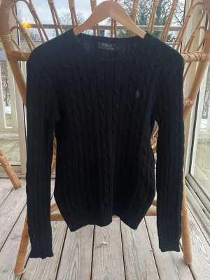 Svart kabelstickad tröja Polo Ralph Lauren - Fet kabelstickad svart tröja från Polo Ralph Lauren med rund halsringning och lila logga. Är en storlek S men passar som en XS. Bara att höra av sig vid frågor. Pris kan diskuteras.