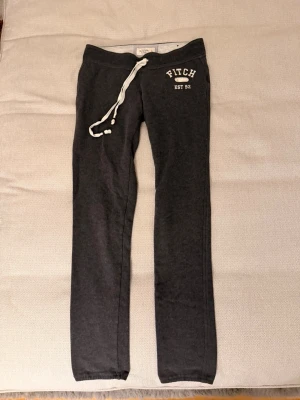 Mörkgrå mjukisbyxor från Abercrombie & fitch - Mörkgrå mjukisbyxor från Abercrombie & fitch med vit snörning i midjan och tryckt logga på vänster höft. Sköna och avslappnade med normal passform, perfekta för chill eller träning. Tillverkade i mjukt bomullsmaterial.
