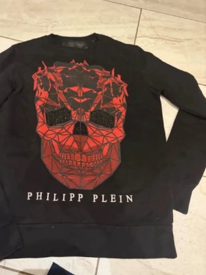 Svart sweatshirt med röd dödskalle - Cool svart sweatshirt Philipp Plein med stor röd geometrisk dödskalle på framsidan och logotypen tryckt under. Tröjan har rund hals och ribbade muddar. Perfekt för dig som gillar streetwear och statement-plagg. Kan diskutera priset 