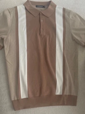 Beige stickad pikétröja J.Lindeberg - Snygg stickad pikétröja från J.Lindeberg i beige och vitt. Tröjan har korta ärmar, krage med två knappar och breda vita ränder framtill. Materialet är tunt och andas, perfekt för en stilren look.