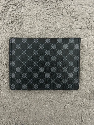 Svart mönstrad plånbok - Stilren svart plånbok med diskret mönster i grått. Plånboken har flera kortfack, ett fönsterfack och stängs med tryckknapp. Tillverkad i tåligt skinn med en klassisk, rektangulär form. Perfekt för dig som gillar snygga detaljer och praktisk design.