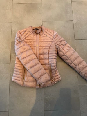 Ljusrosa dunjacka från Esprit - Snygg ljusrosa dunjacka från Esprit med quiltad design och guldfärgade dragkedjor. Jackan har lång ärm, smal passform och står upp lite i kragen. Perfekt för kyliga dagar och ger en stilren look.