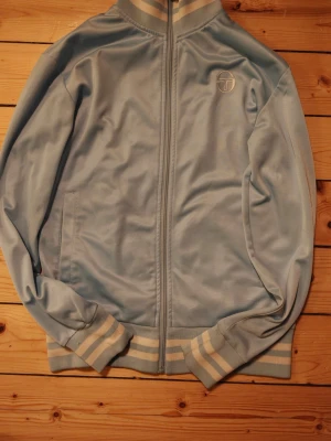 Ljusblå vindjacka från Sergio Tacchini - Ljusblå vindjacka från Sergio Tacchini med dragkedja framtill och broderad logga på bröstet. Jackan har ribbade muddar och krage med vita ränder samt två sidofickor. Tillverkad i mjuk polyester för en sportig och retro look.