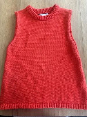Röd/orange stickad väst från Zara. - Säljer en snygg röd/orange stickad väst från Zara med rund halsringning och ribbade kanter. Västen är ärmlös och har en enkel, rak passform som passar perfekt över en skjorta eller t-shirt. Materialet är mjukt och ger en trendig look.