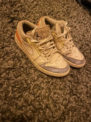 Nike Air Jordan 1 Low beige/orange - Snygga Nike Air Jordan 1 Low sneakers i beige med orange detaljer på hälen och grå partier vid tån. Klassisk siluett med snörning, rund tå och platt sula. Ikonisk Jumpman-logga på plösen. Perfekta för dig som gillar streetwear och vill sticka ut.