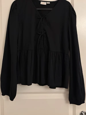 Svart blus med knyt och volanger - Svart blus med v-ringning och två dekorativa knyt framtill. Blusen har långa ärmar med lätt puff och resår vid ärmslut. Nederdelen är volangig för en luftig känsla. Perfekt till jeans eller kjol för en trendig look.