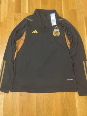 Argentina träningsset från Adidas - Mörkbrunt träningsset från Adidas med guldiga detaljer och AFA-logga. Setet består av en långärmad tröja med halv dragkedja och matchande byxor med tre guldiga ränder längs benen. Perfekt för fotbollsträning eller chill. Materialet är troligtvis polyester.