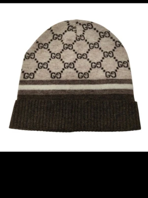Beige och brun mössa från GUCCI - Snygg beige och brun mössa från Gucci med klassiskt GG-mönster över hela ytan. Mössan har bred ribbstickad kant och två kontrasterande ränder. Tillverkad i mjukt stickat material som känns lyxigt och trendigt.Bl a Kashmir.... Riktigt fin o unik.Allt som jag lägger upp är autentiska och har äkthetsbevis med serienummer!!
