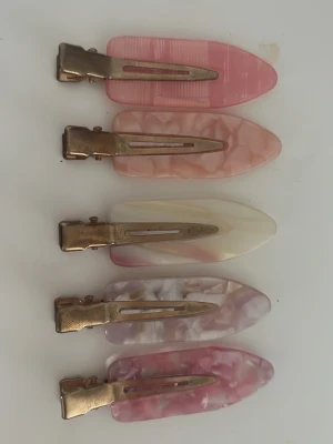 Fem färgglada hårklämmor i pastell - Säljer ett set med fem hårklämmor i olika pastellfärger som rosa, beige och lila. Klämmorna har en avlång, avsmalnande form och är tillverkade i plast med metallspänne. Några har marmorerat mönster och andra är enfärgade. Perfekta för att piffa till frisyren!