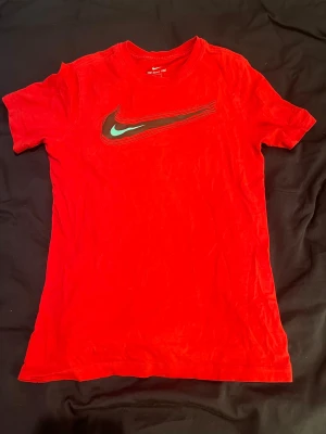  Nike t-shirt - Röd t-shirt från Nike med en stor, snedställd Swoosh-logga i svart och grönt på bröstet. Klassisk rund hals och korta ärmar. Tillverkad i mjuk bomull som känns skön mot huden. Perfekt för en sportig och avslappnad stil.