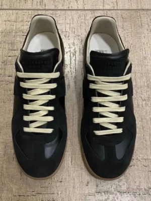 Svarta sneakers från Maison Margiela - Snygga svarta sneakers från Maison Margiela med beige skosnören och klassisk gummisula. Ovandelen är i en mix av mocka och slätt läder, med diskret logotyp på plösen. Säljer för de är för små för min son. Aldrig använda!