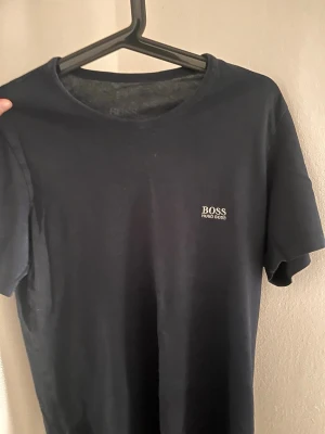 Svart t-shirt från Hugo Boss - Klassisk svart t-shirt från Hugo Boss med diskret logga på bröstet. T-shirten har rund halsringning och korta ärmar, perfekt för en clean och stilren look. Tillverkad i mjuk bomull som känns skön mot huden.
