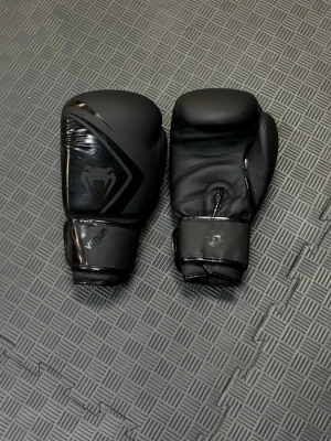 Svarta boxningshandskar från Venum - ||PRISET KAN SÄNKAS VID DIREKT KÖP|| Säljer ett par svarta boxningshandskar från Venum 14 oz. Perfekt för sparing och säck, använt max 5 ggr, nyskick. matt och glansig finish. Handskarna har kardborreknäppning vid handleden och en stilren design med Venum-loggan på ovansidan. Perfekta för boxning eller träning.