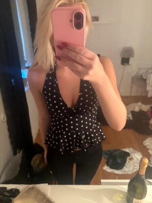 Svart prickig omlottopp med volang - Snygg svart omlottopp med vita prickar och volangkant nedtill. Toppen har djup v-ringning och breda axelband som knyts i nacken. Perfekt för dig som vill sticka ut lite extra med ett lekfullt mönster.