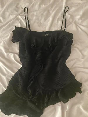 Svart volangig linnetopp från Bik Bok - Supersnygg svart linnetopp från Bik Bok med tunna axelband och volangdetaljer framtill. Toppen har en plisserad struktur och är perfekt för dig som vill ha en trendig och feminin look. Passar till både jeans och kjol.