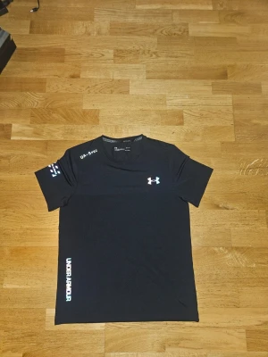 Svart träningströja från Under Armour - Snygg svart träningströja från Under Armour med korta ärmar och logga på bröstet. Tröjan har tryck på ena ärmen och längs sidan, samt UA-Spec text vid axeln. Tillverkad i ett lätt och snabbtorkande funktionsmaterial som passar perfekt till träning.