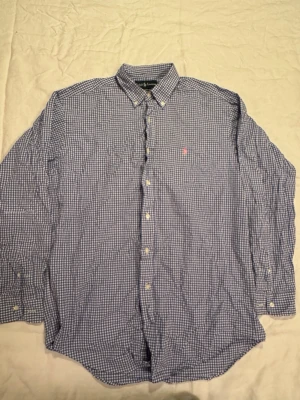 Blårutig skjorta från Ralph Lauren - Klassisk blårutig skjorta från Ralph Lauren i bomull. Skjortan har lång ärm, button-down krage och vita knappar framtill. Liten broderad logga i rött på bröstet. Perfekt för en clean och stilren look.