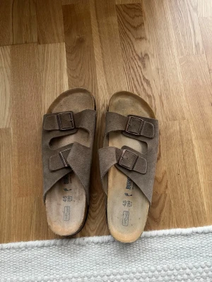 Beige mockasandaler från Birkenstock - Snygga sandaler från Birkenstock i beige mocka med två breda remmar och justerbara metallspännen. Klassisk design med platt sula och rund tå, perfekt för sommarens alla dagar. Märket är ingraverat på spännena för extra stilpoäng. Finns ingen kartong till! DU VET VARFÖR DE ÄR BILLIGA