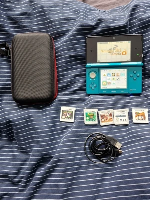Blå Nintendo 3DS med spel och tillbehör - Säljer en blå Nintendo 3DS med fem spel, laddkabel och ett svart skyddsfodral. Konsolen har dubbla skärmar, klassisk vikbar design och tydliga knappar. Perfekt för dig som gillar portabel gaming och vill ha allt i ett paket.