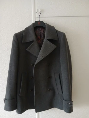 Mörkgrå skepparkavaj från Oscar Jacobson - Stilren mörkgrå skepparkavaj från Oscar Jacobson med dubbelknäppning och bred krage. Kavajen har två sidofickor, dekorativa knappar vid ärmslut och ett paisleymönstrat innerfoder. Tillverkad i ull för en klassisk look.