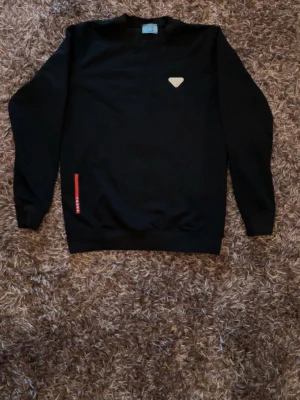 Svart sweatshirt från Prada - Säljer en svart sweatshirt från Prada med rund halsringning och långa ärmar. Tröjan har en liten vit logotyp på bröstet och en röd detalj med Prada-tryck vid nederkanten. Perfekt för en clean och stilren look.