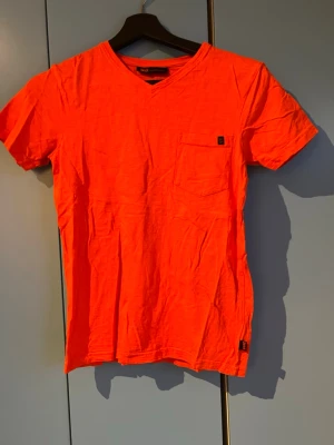 Orange t-shirt med v-ringning - Snygg orange t-shirt från Solid med v-ringad hals och liten bröstficka. T-shirten är kortärmad och har en enkel, stilren design som passar perfekt till jeans eller shorts. 