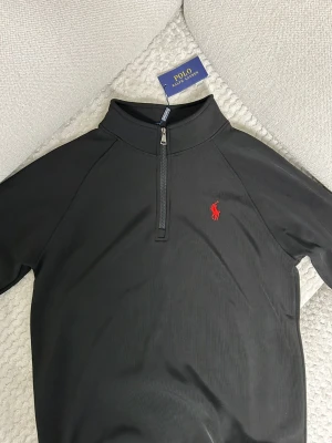 Svart half-zip tröja Polo Ralph Lauren - Svart långärmad half-zip tröja från Polo Ralph Lauren med röd broderad logga på bröstet. Tröjan har hög krage och dragkedja framtill. Tillverkad i mjukt material med en sportig och stilren look.