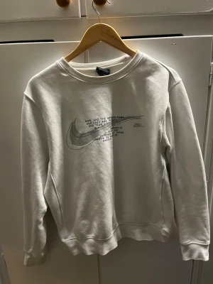 Vit sweatshirt från Nike med tryck - Säljer en vit sweatshirt från Nike med klassiskt Swoosh-tryck och text på bröstet. Tröjan har rund halsringning, långa ärmar och ribbade muddar. Perfekt för en avslappnad och sportig stil. Passar som s/m