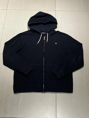 Marinblå hoodie från Morris - Snygg marinblå hoodie från Morris med dragkedja framtill och vit dragsko med läderdetaljer. Klassisk passform, huva och liten broderad logga på bröstet. Perfekt för en avslappnad och stilren look. Tillverkad i mjuk bomull.