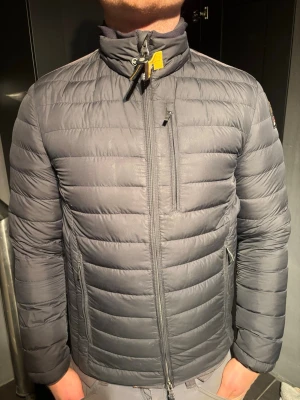 Mörkgrå dunjacka från Parajumpers - Snygg mörkgrå dunjacka från Parajumpers med hög krage, dragkedja framtill och flera fickor. Jackan har quiltad design och en gul detalj vid halsen. Perfekt för kalla dagar och riktigt stilren look. Den är i helt nytt skick och äkta.