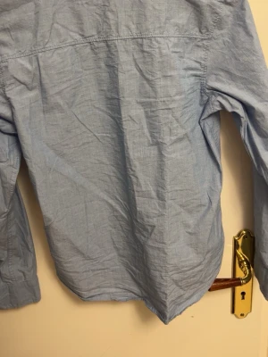 Ljusblå klassisk skjorta i bomull - En stilren ljusblå skjorta med lång ärm och klassisk krage. Skjortan är tillverkad i mjuk bomull och har en enkel, tidlös design utan mönster eller extra detaljer. Perfekt för dig som gillar en clean och avslappnad look.