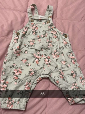 Blommig hängselbyxa från Newbie - Supersöt hängselbyxa från Newbie i ljusgrått med rosa och grönt blommönster. Byxan har knappar i trälook på axelbanden och sidorna samt resår vid bensluten. Perfekt för dig som gillar gulliga och bekväma plagg till de minsta.