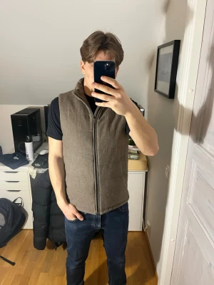 Brun ullväst med dragkedja från John Henric  - Snygg brun väst i ullmaterial med hög krage och hel dragkedja framtill. Västen har två diskreta fickor på sidorna och även två innerfickor och alla är magnetiska. Strl S