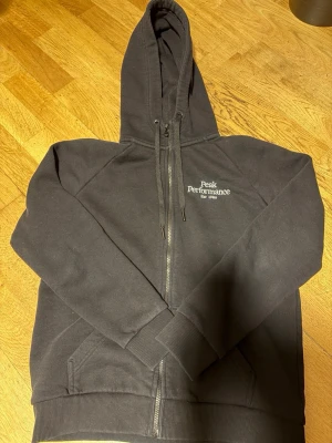 Svart zip-hoodie Peak Performance - Snygg svart hoodie med dragkedja från Peak Performance. Klassisk design med huva och dragsko, två fickor framtill och logga broderad på bröstet. Tillverkad i mjukt bomullsmaterial som känns skönt mot huden. Perfekt för en avslappnad och sportig stil.