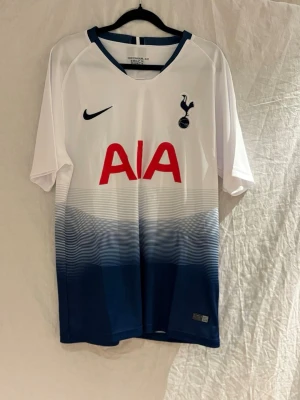 Tottenham Dele #20 - Säljer en Tottenham Hotspur matchtröja från med Dele och nummer 20 på ryggen. Tröjan är vit upptill och övergår i mörkblått nertill med gradient-effekt. Stor röd AIA-logga på bröstet och klubbmärke på vänster sida. Perfekt för fotbollsfans!